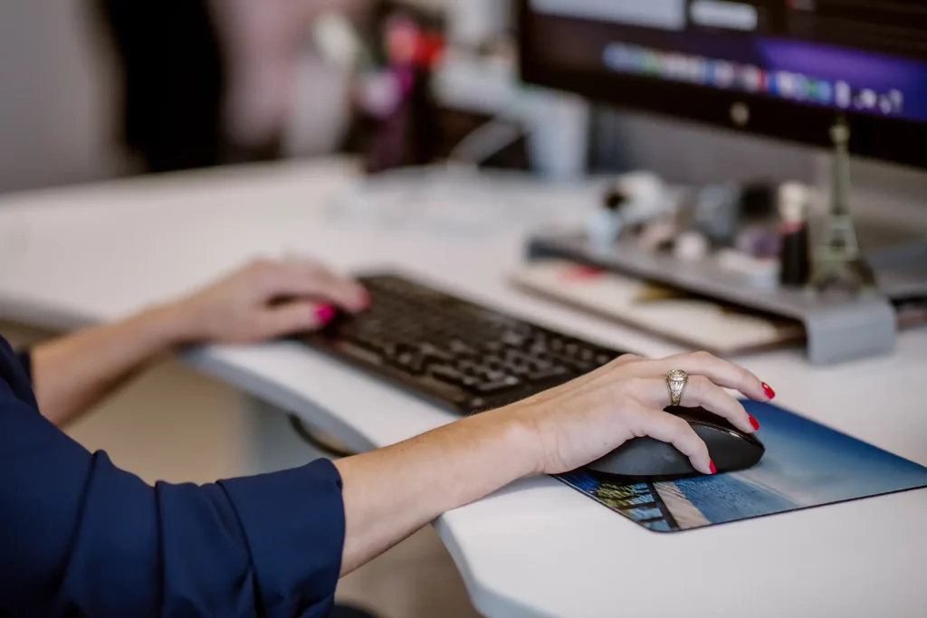 A woman with red fingernails types on a keyboard and holds a computer mouse.