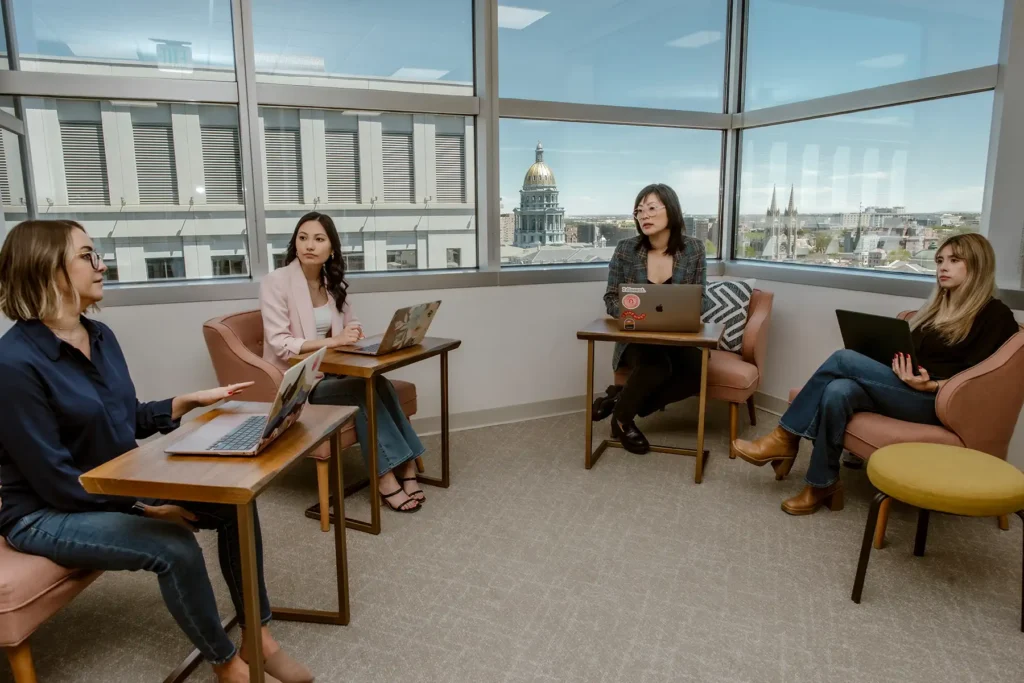 Four professional women with laptops have a meeting in front of a high rise window.