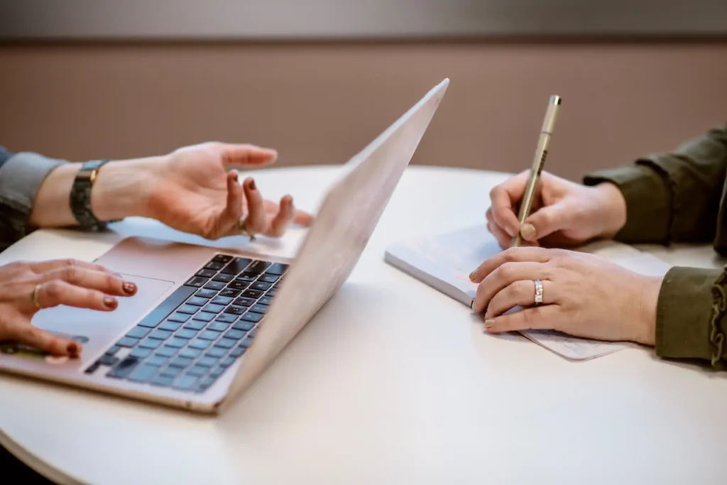 A woman with a laptop sits at a desk with a woman writing in a notebook.