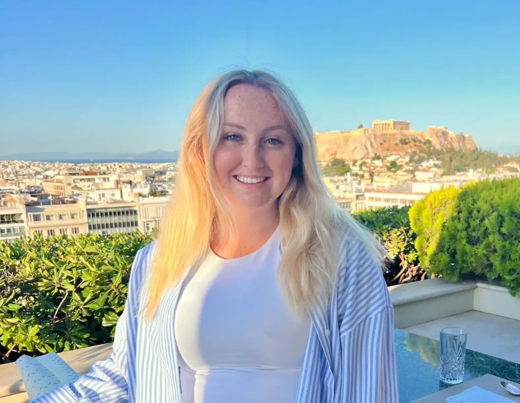 Emily smiling on a balcony on a sunny day with a city behind her.