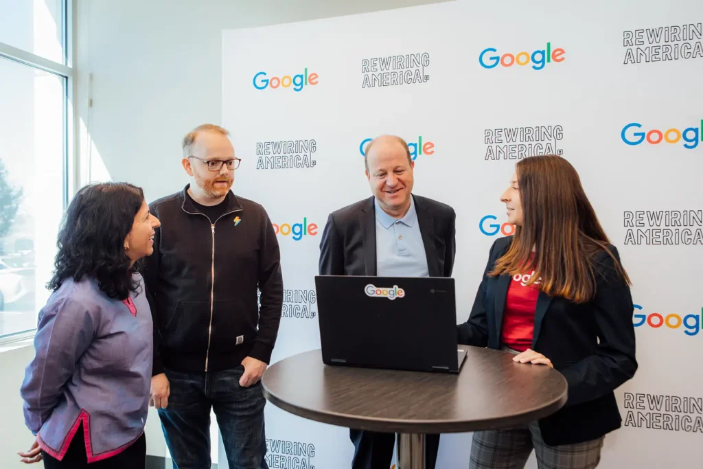Jared Polis speaking with 3 others in front of a backdrop that has Google and Rewiring America logos on it