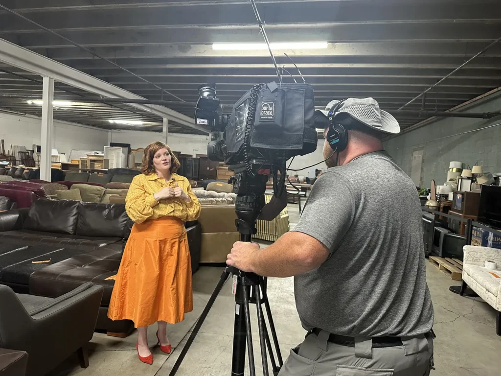 A client from Furnishing Hope stands in front of a camera while she is interviewed