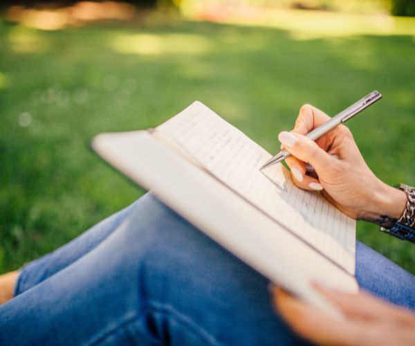 A person writes in a notebook while sitting in a lawn of grass
