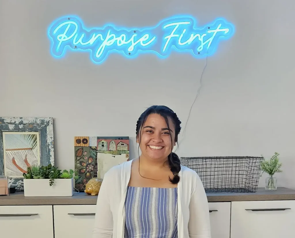 Ana, design intern, posing in front of a lit up sign that reads "Purpose First"