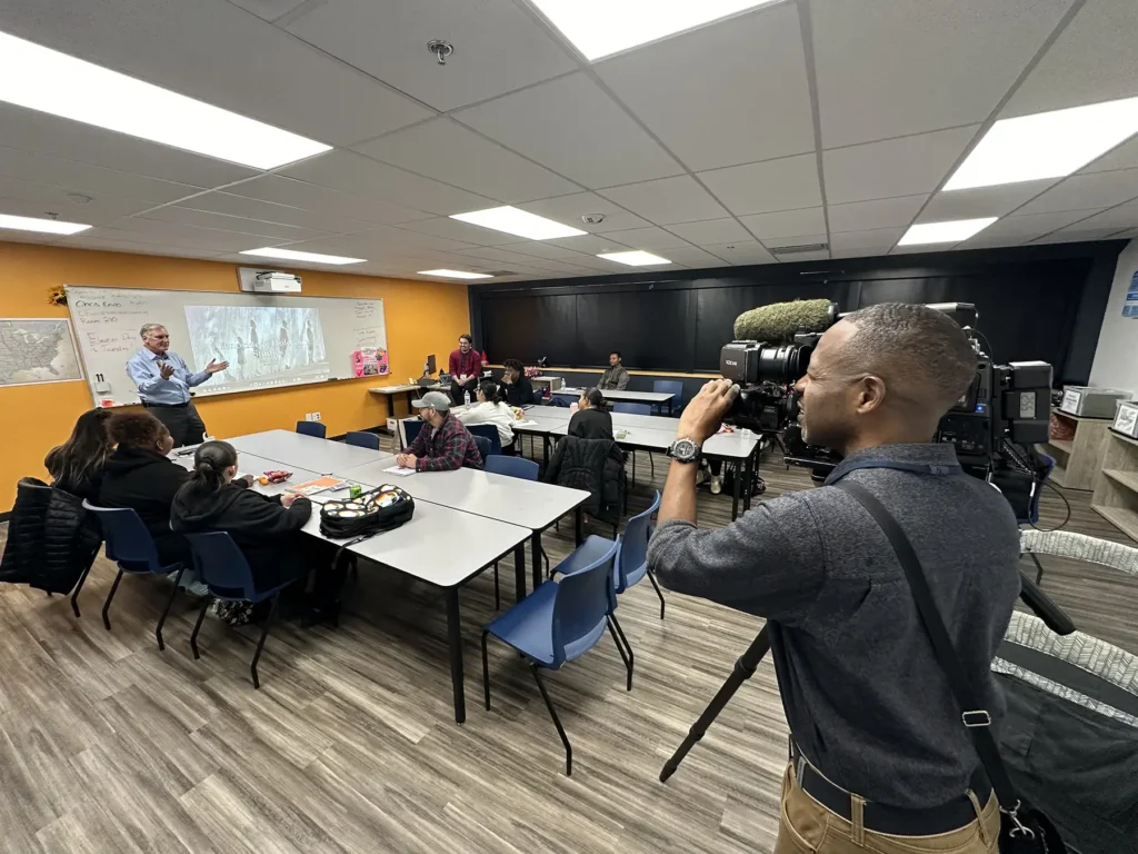 A man operating a camera points it at another man teaching something in a classroom.