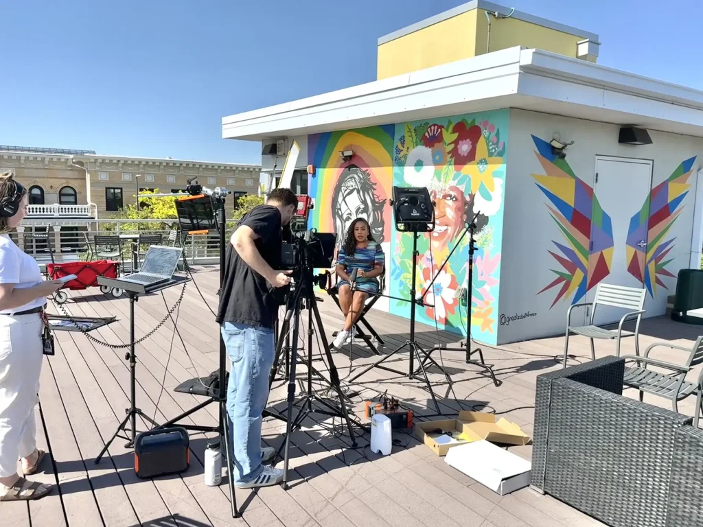 A woman is being interviewed by a camera crew in front of a mural of Marsha P. Johnson and other LGBTQ+ art