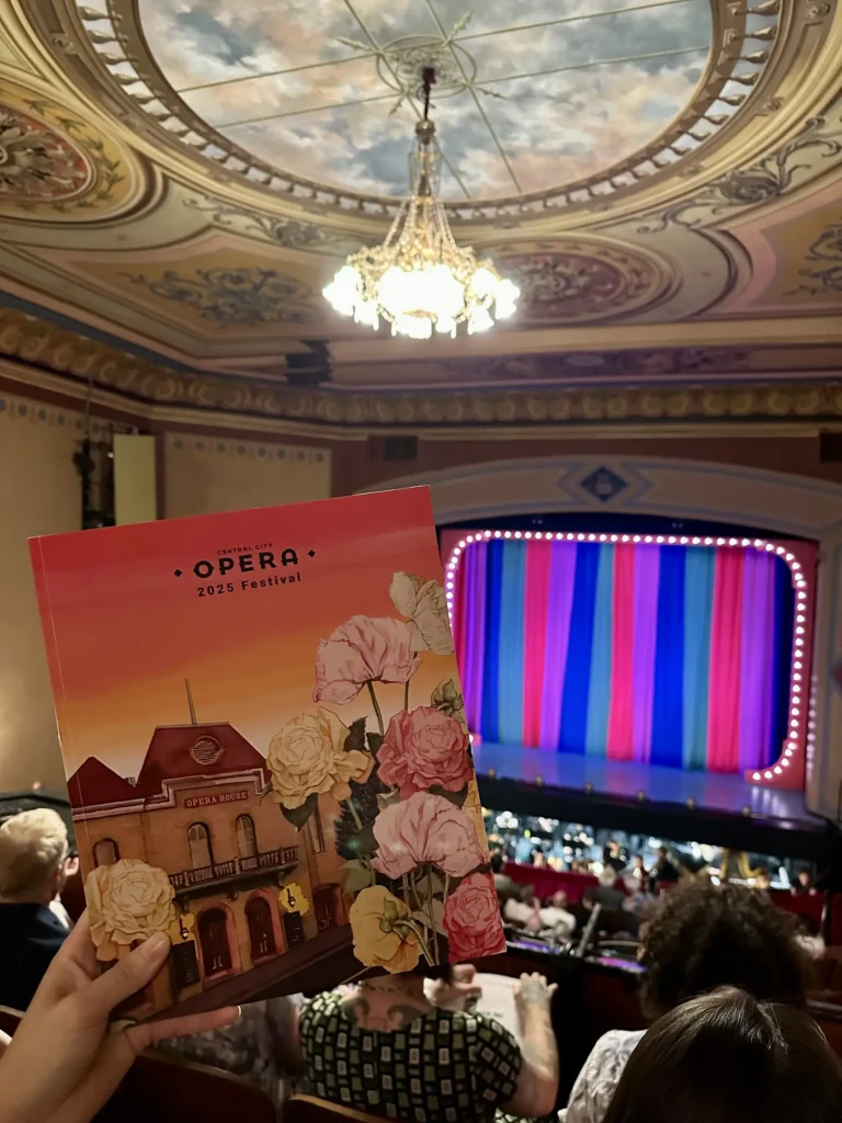 A designed festival program book with flowers and an opera house on the cover being held in front of an actual opera house auditorium