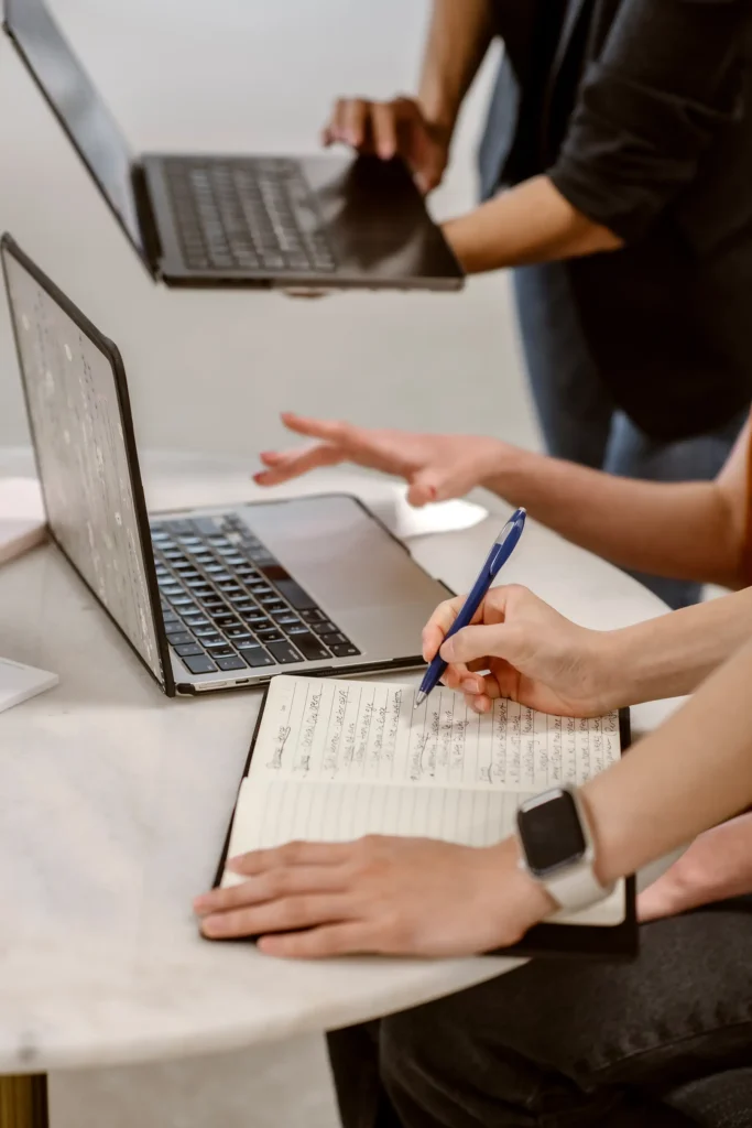 A group of women discuss something in front of laptops and a notebook